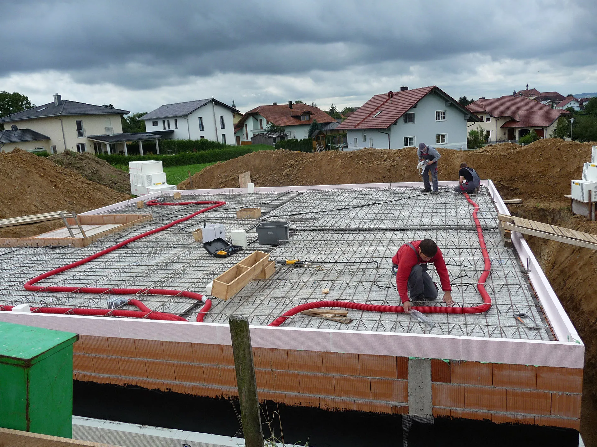 Mehrere Arbeiter der Neudorfer GmbH installieren ein Lüftungssystem im Fundament eines Neubaus, wobei auffällig rote Lüftungsleitungen über einem Stahlgittermuster verlaufen. 
