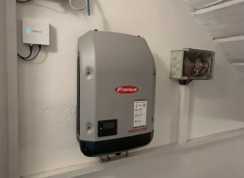 Ein silberner Wechselrichter der Marke Fronius ist an einer weißen Wand unter einer Dachschräge montiert.