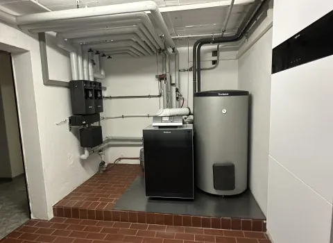 Im Heizungskeller ist eine Ölhybridanlage installiert, bestehend aus einem grauen Speicherbehälter und einem danebenstehenden schwarzen Heizgerät von Buderus.