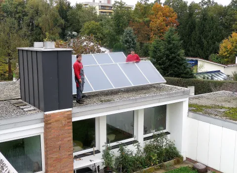Zwei Anlagenmechaniker der Neudorfer GmbH installieren eine große thermische Solaranlage auf dem Flachdach eines modernen Wohnhauses.