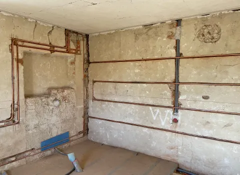 Ein Wanddetail zeigt offenliegende Kupferrohre, die horizontal und vertikal durch Schlitze in einer Altbauwand verlaufen. 