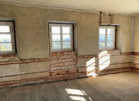 In einem renovierungsbedürftigen Raum sind Wandheizungsrohre sichtbar in das Mauerwerk unterhalb von drei Fenstern integriert.