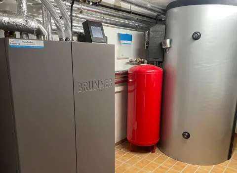 Ein kompakter Heizraum zeigt eine moderne BRUNNER-Heizungsanlage mit integriertem Steuerdisplay, daneben ein silberner Pufferspeicher und ein leuchtend rotes Ausdehnungsgefäß.