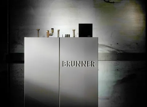 Ein BRUNNER Pelletskessel steht vor einer metallischen Wand mit industrieller Anmutung. 