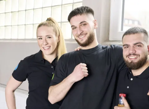 Drei junge Auszubildende Anlagenmechaniker posieren in Arbeitskleidung vor dem Werkstattsfenster.