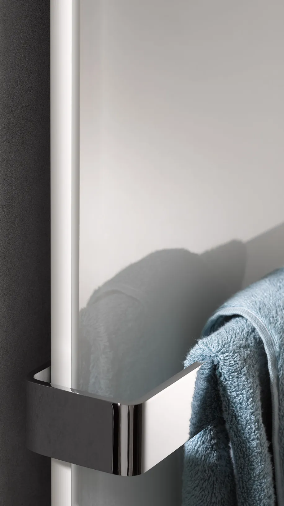 Detailaufnahme eines Glasheizkörpers mit verchromter Halterung, an der ein blaues Handtuch hängt, vor einer dunklen Wand.