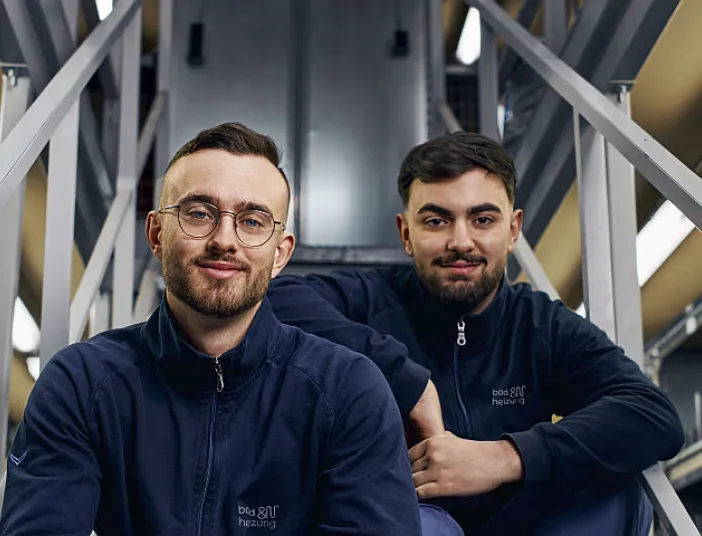 Zwei junge Männer in Arbeitskleidung von bad und heizung sitzen auf einer Metalltreppe in einem Lagerraum, umgeben von Regalen voller Ersatzteile und Verpackungen. 
