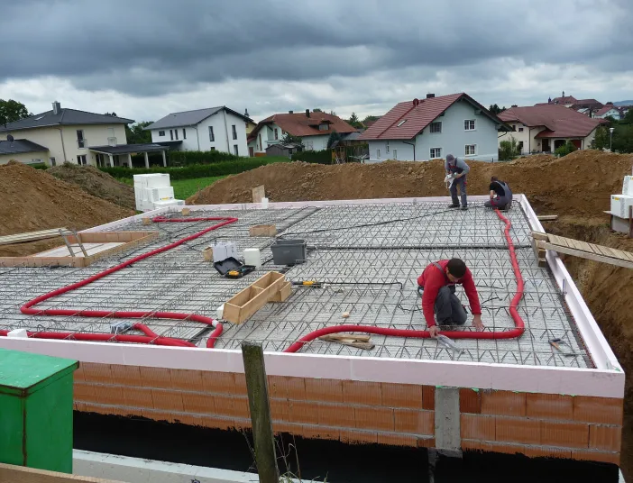 Mehrere Arbeiter der Neudorfer GmbH installieren ein Lüftungssystem im Fundament eines Neubaus, wobei auffällig rote Lüftungsleitungen über einem Stahlgittermuster verlaufen. 