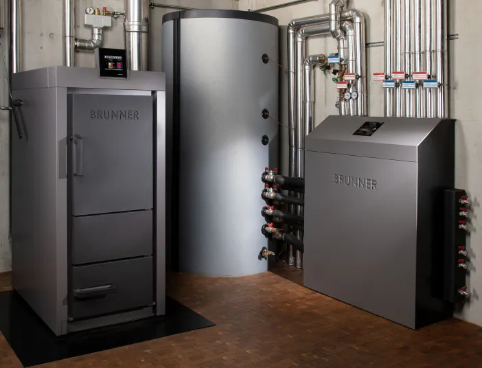 In einem modernen Heizraum steht eine Stückholzanlage von Brunner, bestehend aus einem großen Brennkessel, einem Pufferspeicher und einer Systemeinheit. 